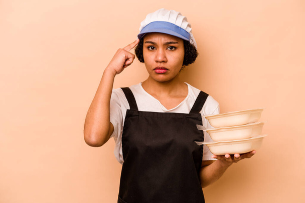 Young cook volunteer African American woman isolated on beige background pointing temple with finger, thinking, focused on a task. - Foto, imagen