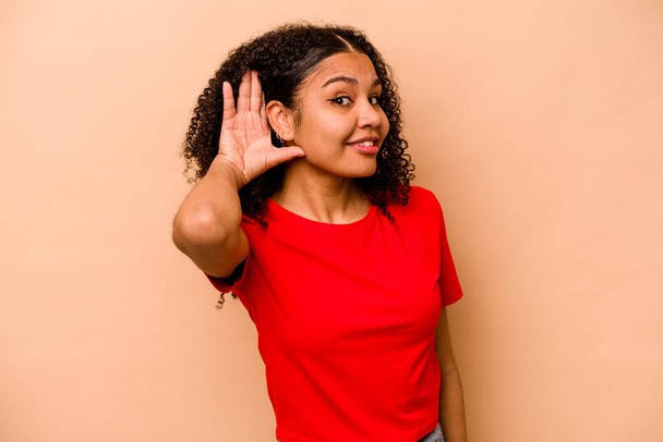 Giovane donna afroamericana isolata su sfondo beige cercando di ascoltare un pettegolezzo. - Foto, immagini