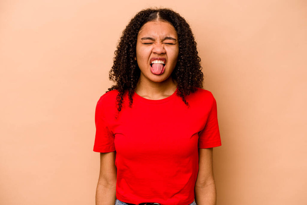 Joven afroamericana americana aislada sobre fondo beige divertida y amigable sobresaliendo lengua. - Foto, Imagen