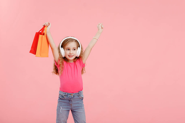 adorable niña en los auriculares está sosteniendo bolsas de compras en las manos por encima de la cabeza aislado sobre fondo rosa. concepto de compra. - Foto, Imagen