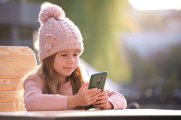 Portrait of cute little child girl in pink hat sitting alone at street cafe playing game on sellphone - Foto, immagini