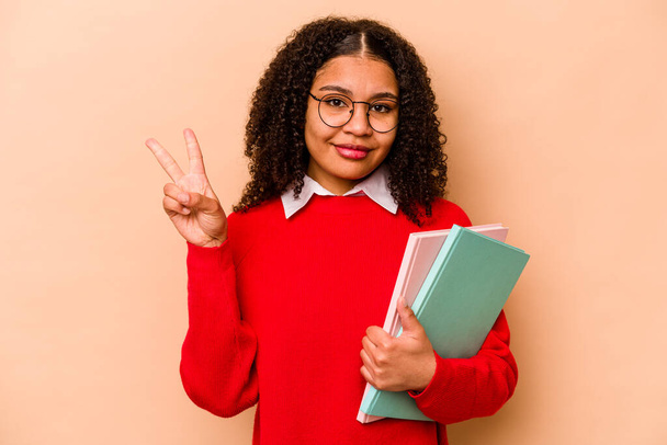Joven estudiante afroamericana aislada sobre fondo beige mostrando el número dos con los dedos. - Foto, Imagen