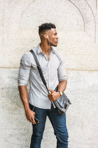Um homem a caminho do trabalho. Vestindo camisa cinza, jeans, relógio de pulso, carregando bolsa de couro de ombro, um cara afro-americano de pé na parede na rua, ouvindo música com fone de ouvido e telefone celular - Foto, Imagem