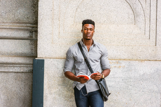 Moderni opiskelija New Yorkissa. Harmaa paita, rannekello, olkalaukku, afroamerikkalainen mies seisomassa kadulla kuuntelemassa musiikkia kuulokkeilla, lukemassa punaista kirjaa. - Valokuva, kuva