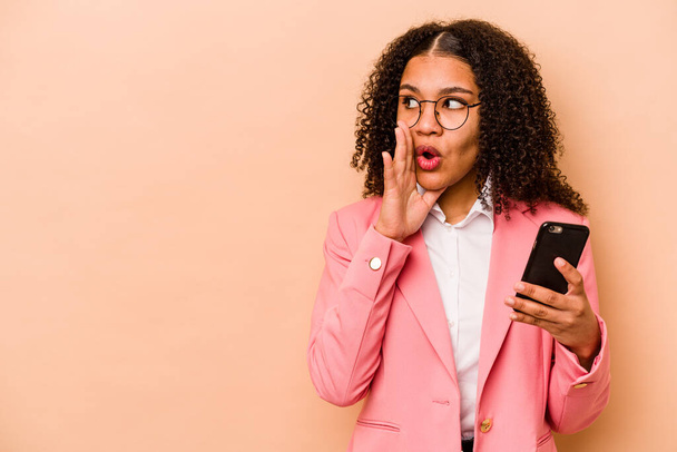 Young African American woman holding mobile phone isolated on beige background is saying a secret hot braking news and looking aside - Foto, Bild