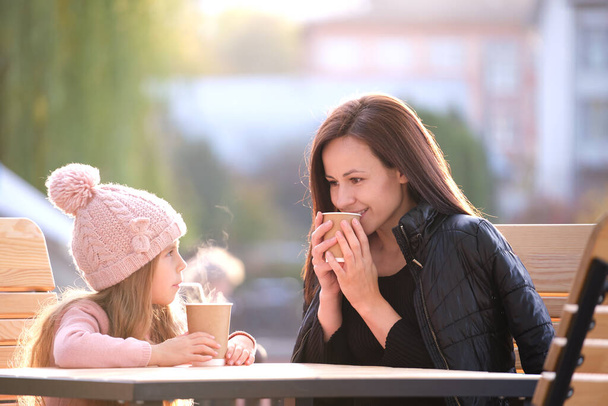 Young mother and her child daughter having good time together sitting at street cafe with hot drinks on sunny fall day. Happiness in family relations concept - Photo, Image