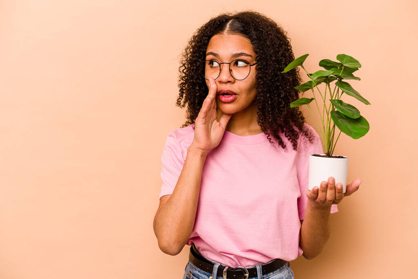 Junge Afroamerikanerin hält eine Pflanze isoliert auf beigem Hintergrund und erzählt eine geheime heiße Bremsnachricht und schaut zur Seite - Foto, Bild