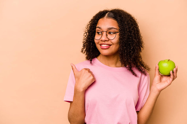 Young African American woman holding an apple isolated on beige background points with thumb finger away, laughing and carefree. - Valokuva, kuva