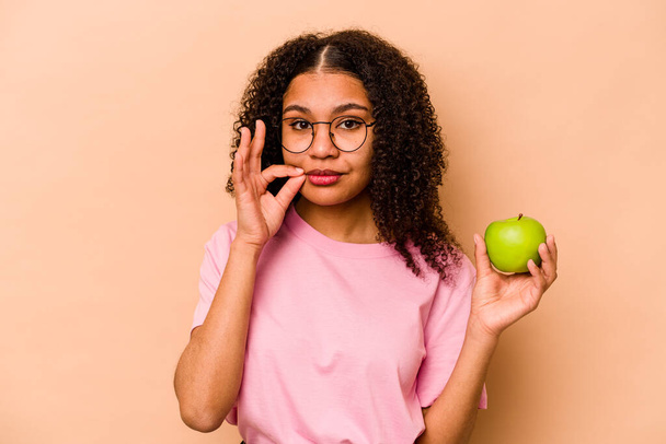 Молодая афроамериканка держит яблоко, изолированное на бежевом фоне с пальцами на губах, хранящими секрет. - Фото, изображение