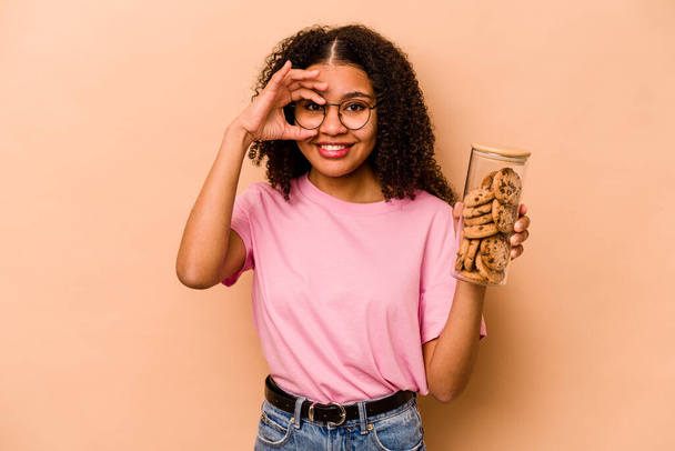 Νεαρή Αφροαμερικανή γυναίκα κρατώντας ένα βάζο cookies απομονωμένο σε μπεζ φόντο ενθουσιασμένοι κρατώντας ok χειρονομία στο μάτι. - Φωτογραφία, εικόνα