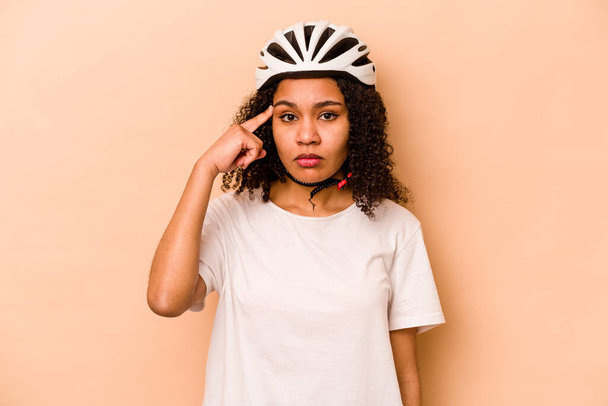 Молода іспаномовна жінка в шолом-велосипеді ізольована на синьому фоні, вказуючи храм пальцем, думаючи, зосереджена на завданні
. - Фото, зображення