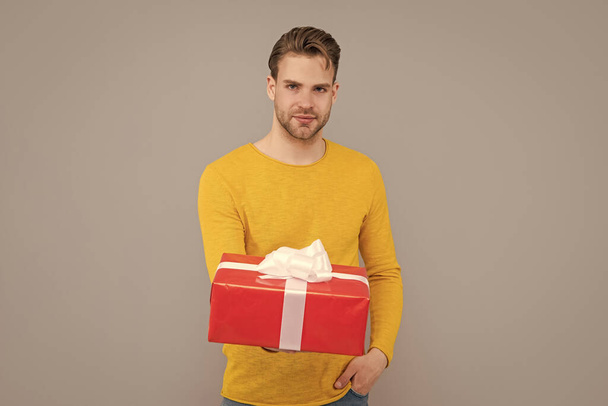 Knappe vent in gele trui houden geschenkdoos grijze achtergrond, gifting - Foto, afbeelding
