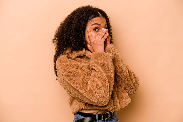 Mladý africký Američan žena izolovaný na béžové pozadí blikat přes prsty vyděšený a nervózní. - Fotografie, Obrázek
