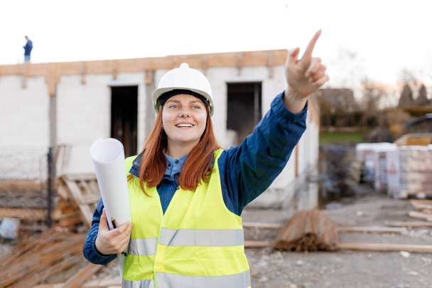 Architect redhead woman wearing contractor white helmet and safety green vest pointing finger. Worker showing something at new building site, concept of planning new project - Foto, imagen