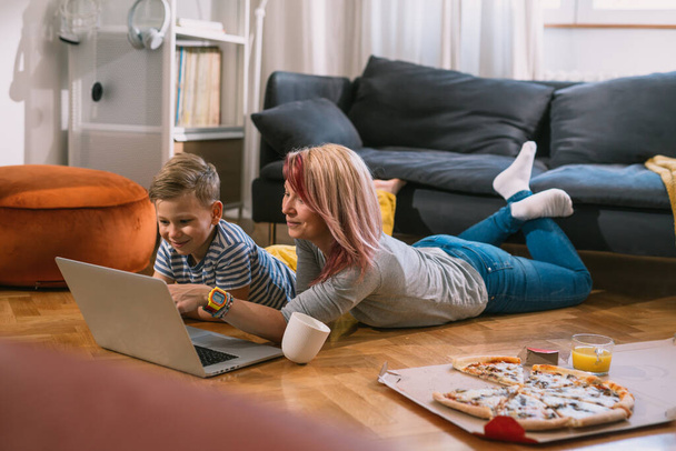 mother and son laying on floor, eating pizza and watching movie on laptop computer in they living room - 写真・画像