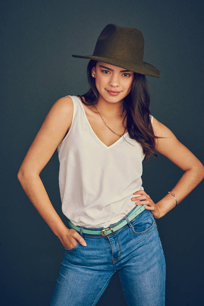 She wears a hat like a pro. Studio shot of an attractive young woman wearing a hat and posing while standing against a dark background. - Foto, imagen