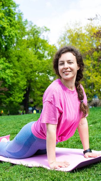 Young athletic woman practicing yoga workout, smiling looking at camera on green grass in city garden park outdoors in summer day, wearing pink t-shirt. Active life, sport, healthy lifestyle.Vertical - Photo, Image