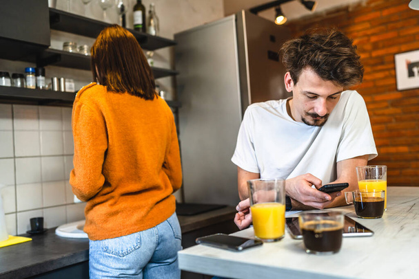 Two people young couple adult man and woman husband and wife or boyfriend with girlfriend in the kitchen in the morning with coffee juice mobile phone and digital tablet talking and chat daily routine - Photo, Image