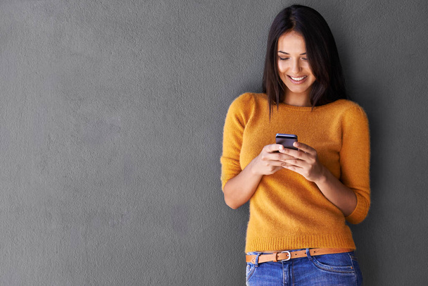 Strahlend und sprudelnd mit einigen guten Nachrichten. Eine attraktive junge Frau benutzt ein Handy, während sie vor einer grauen Wand steht. - Foto, Bild