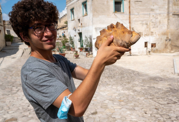 Matera, Basilicata, Italia Agosto de 2021. Un joven turista sostiene una forma del pan típico de la ciudad. Hecho de sémola de trigo duro, tiene un color amarillento. El centro histórico en el fondo. - Foto, imagen