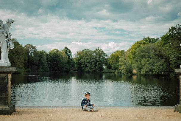 A little boy sits by the river on a sunny day - Zdjęcie, obraz