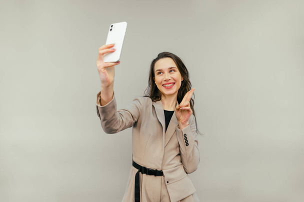 Joyful dame in een jas staat op een beige achtergrond met een glimlach op haar gezicht neemt een selfie en toont een gebaar van vrede, poseren in de smartphone camera. - Foto, afbeelding