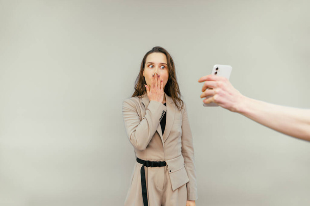 Shocked business woman in a suit looks at the smartphone screen in someone else's hand and holds her face in surprise, isolated on a beige background. - Photo, image