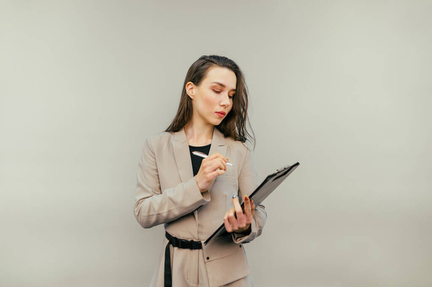 Attractive woman in a suit stands on a beige background with a tablet in hand and writes with a pen on paper with a serious face. Businesswoman makes notes. - Фото, зображення