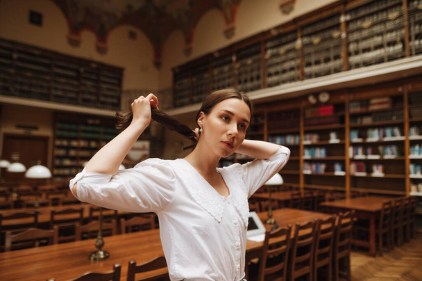 Atrakcyjna dziewczyna w białej bluzce pozuje do kamery w starej, atmosferycznej bibliotece publicznej. - Zdjęcie, obraz