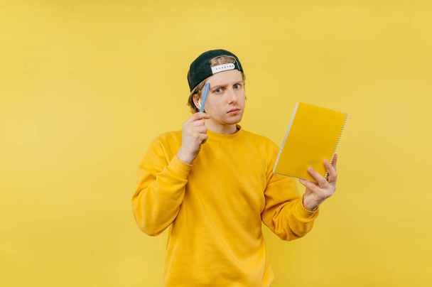 Portrait of a pensive guy in a cap with a notebook in his hand and a pen standing on a yellow background and looking at the camera and thinking, pointing to his temple - Foto, immagini