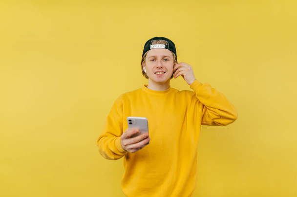 Positive young man in a cap and casual clothes stands on a yellow background with a smartphone in his hands and looks at the camera with a smile on his face, listens to music on headphones - Zdjęcie, obraz