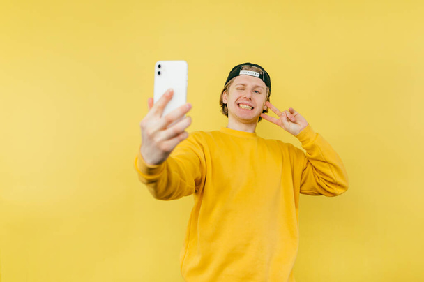 Positive guy in a cap and yellow sweatshirt takes a selfie on camera and smartphone and shows a gesture of peace and winks - Fotó, kép