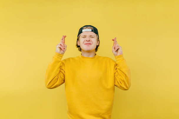 Aranyos srác sapkában simogatja a vágyat keresztbe teszi az ujjait a sárga háttér és mosolygós, elszigetelt sárga háttér. - Fotó, kép