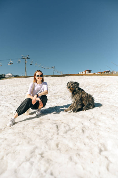 Positive female hiker in casual clothes sitting on snow in mountains and posing at camera with smile on face near dog. - Фото, изображение