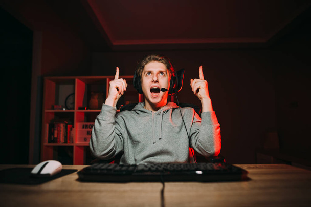 alegre chico gamer en auriculares jugando juegos en línea en casa en el ordenador y apuntando pulgares hacia arriba en el espacio de copia - Foto, Imagen