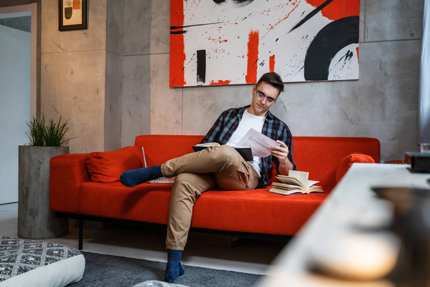 Yksi mies nuori aikuinen valkoihoinen mies istuu sohvalla yksin kotona lukiessaan ja opiskellessaan tai analysoidessaan raporttia tai tenttiä kirjan ja paperin asiakirjoista oikeat ihmiset kopioivat tilaa - Valokuva, kuva