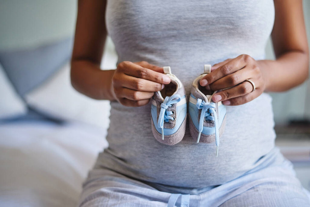 Cute and comfy, just perfect for my little boy. Cropped shot of a pregnant woman holding a pair of blue baby shoes in front of her belly. - Foto, imagen