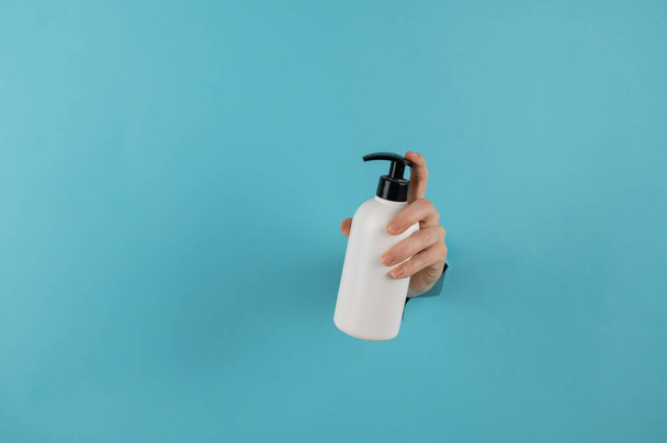 A womans hand sticks out of a hole in a blue paper background and holds a skin lotion pump. - Foto, afbeelding