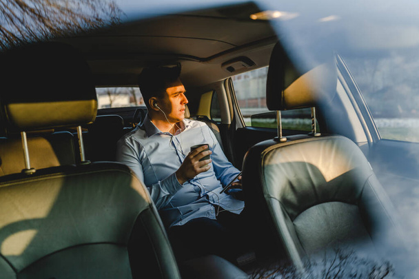 One man young adult male sitting on the back sit of the car wearing shirt holding a cup with coffee or drink looking to the side businessman manager going to work real people copy space - Foto, Imagen