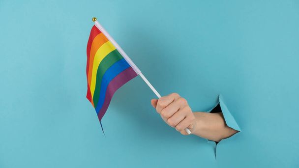 A womans hand sticks out of a hole in a blue paper background and holds an lgbt flag. - Zdjęcie, obraz