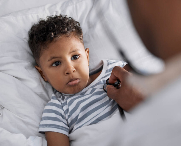 Das tut weh. Aufnahme eines entzückenden kleinen Jungen, der im Bett liegt und sich krank fühlt, während ein Kinderarzt ein Stethoskop benutzt. - Foto, Bild