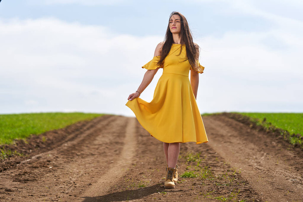 Young woman in a yellow dress on a dirt road among wheat fields - Foto, afbeelding