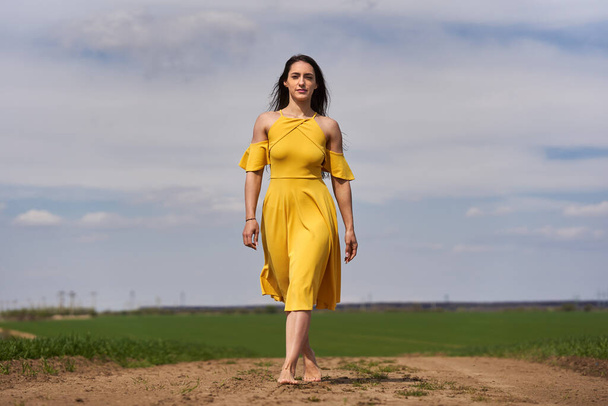 Barefoot young woman in a yellow dress on a dirt road among wheat fields - Fotoğraf, Görsel