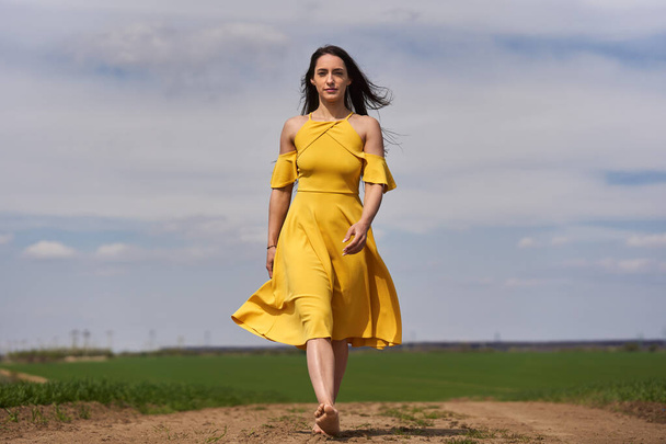 Barefoot young woman in a yellow dress on a dirt road among wheat fields - Fotoğraf, Görsel