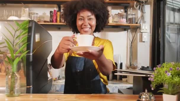 African American female barista in looks at camera, offers cup of coffee to customer with cheerful smile, happy service works in casual restaurant cafe, young small business startup entrepreneur. - Materiał filmowy, wideo