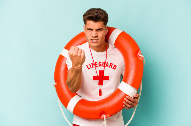 Young caucasian lifeguard isolated on blue background showing fist to camera, aggressive facial expression. - Foto, Imagem