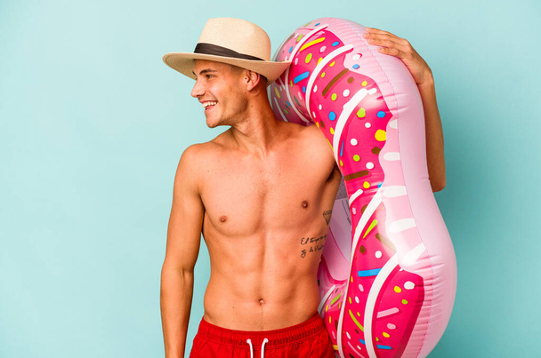 Young caucasian man holding an inflatable donut isolated on blue background looks aside smiling, cheerful and pleasant. - 写真・画像