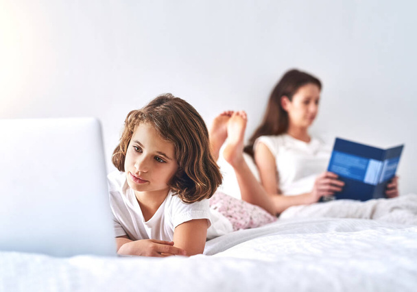He ovat kateellisia omassa maailmassaan. rajattu kuva nuori tyttö käyttää kannettavaa tietokonetta, kun hänen äitinsä lukee kirjan taustalla. - Valokuva, kuva