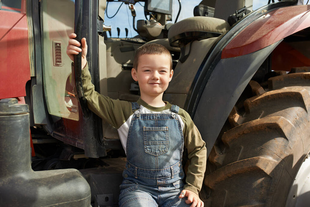 Ilyen nagynak kell lenned, hogy ezt meglovagold. Egy fiatal fiú portréja, aki egy traktor előtt ül.. - Fotó, kép