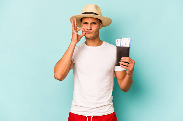 Young caucasian traveler man holding passport isolated on blue background with fingers on lips keeping a secret. - 写真・画像